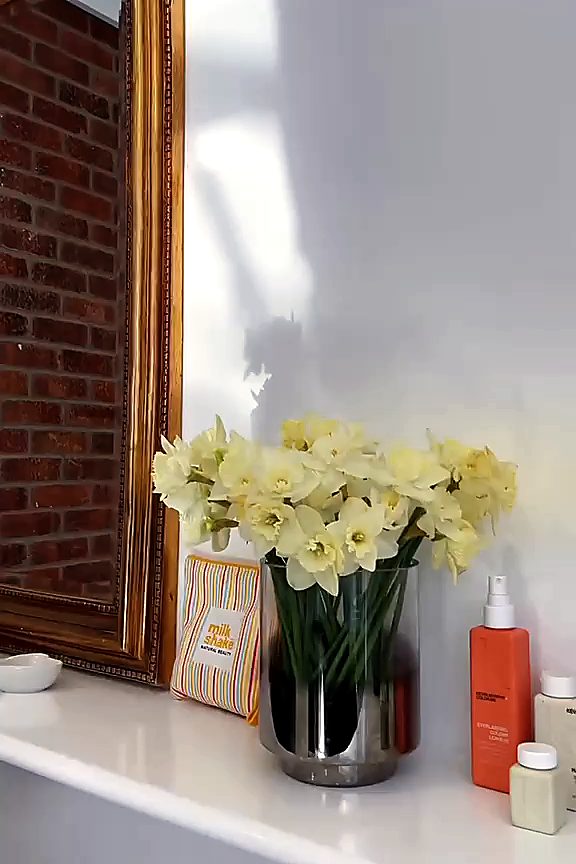 Daffodils in a steel vase on the salon shelf, beside a gold-framed mirror reflecting exposed brick, with sunlight across the wall.